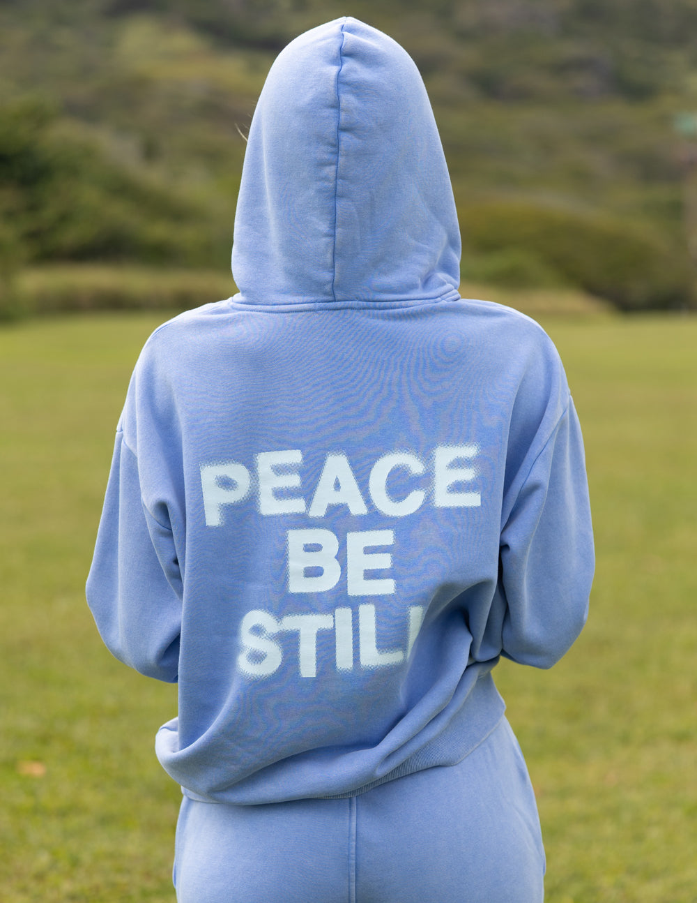 Light blue unisex hoodie with hood up and large “PEACE BE STILL” back print