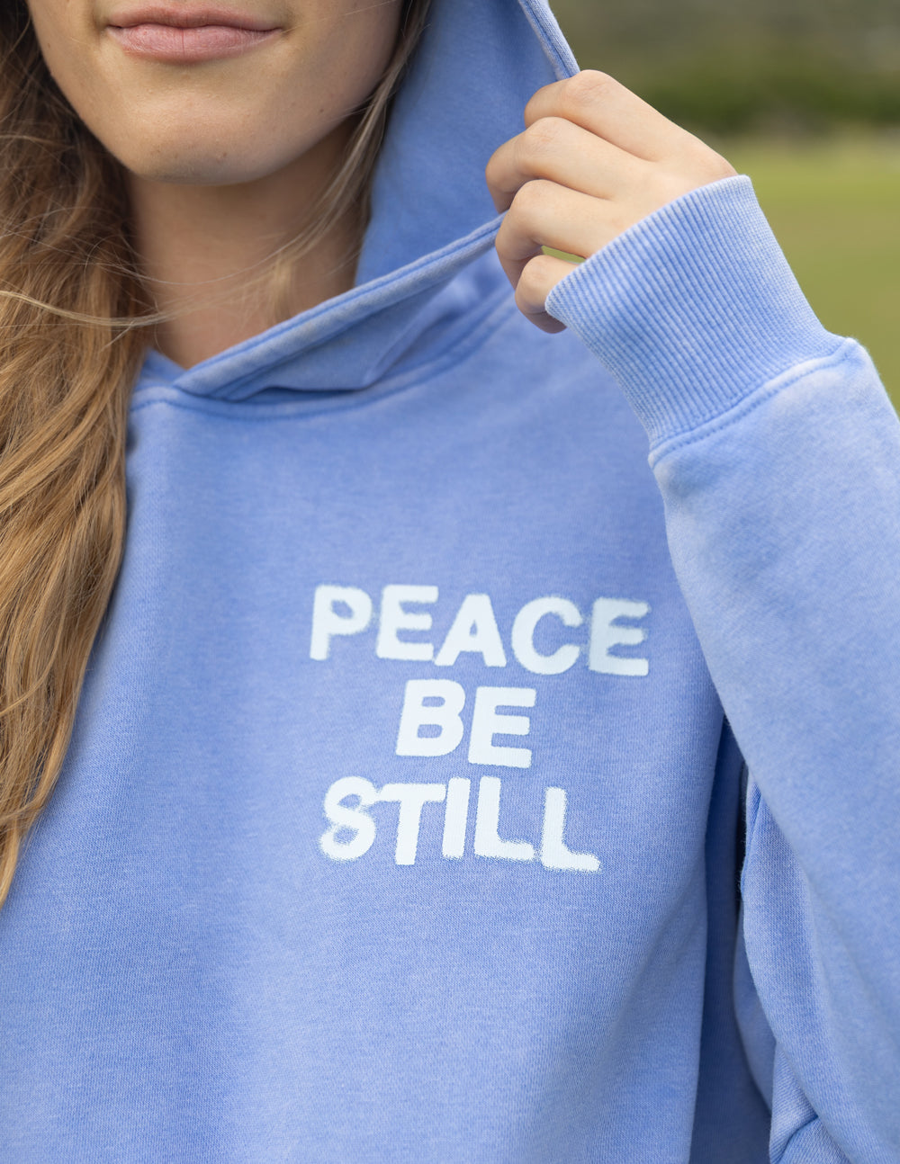 Light blue hoodie with “PEACE BE STILL” front graphic and pullover hood