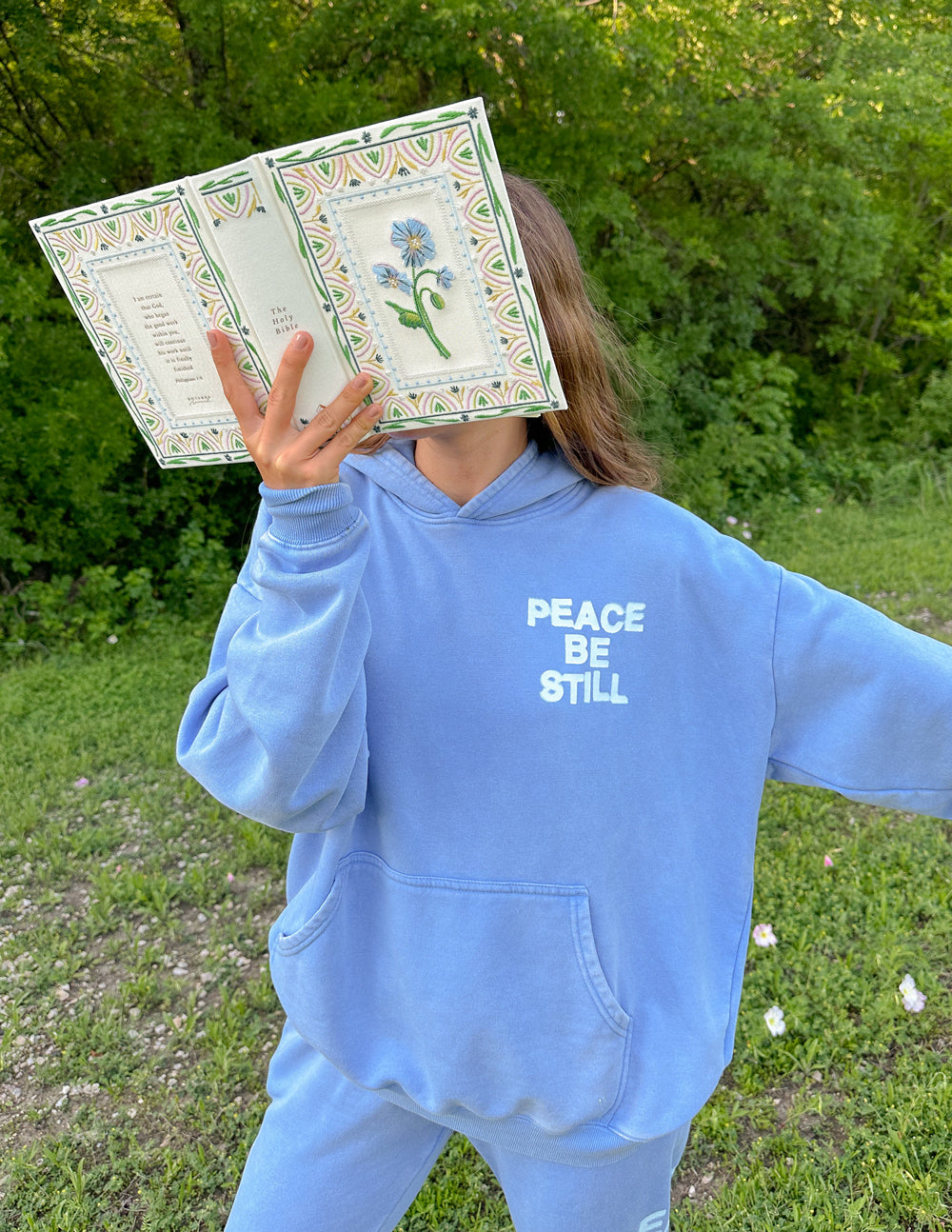 Light blue hoodie with “PEACE BE STILL” chest graphic and front pocket