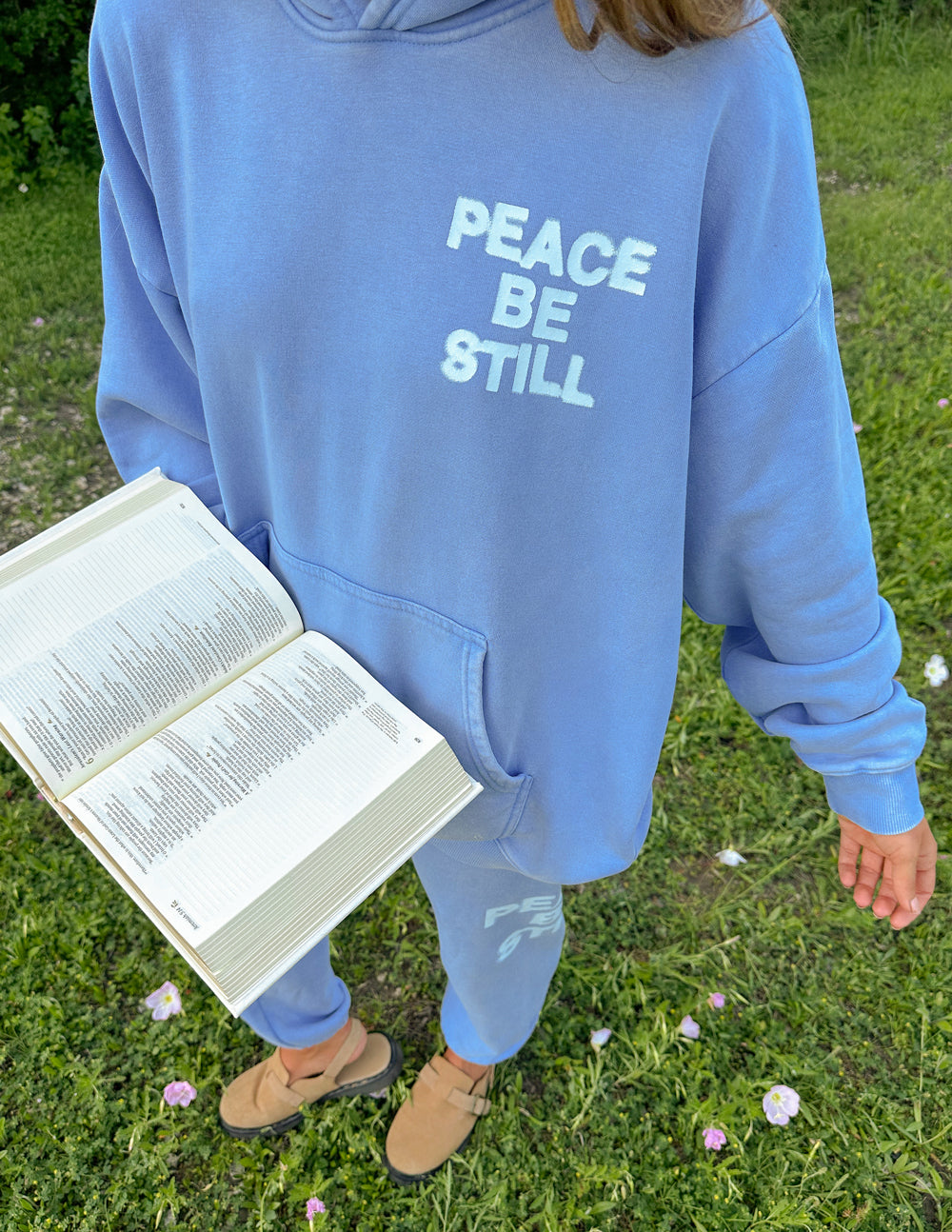 Light blue hoodie front with “PEACE BE STILL” graphic and front pocket