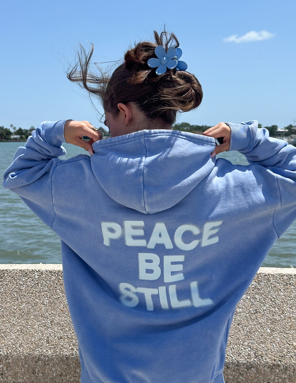 Light blue unisex hoodie with hood down and “PEACE BE STILL” back graphic print