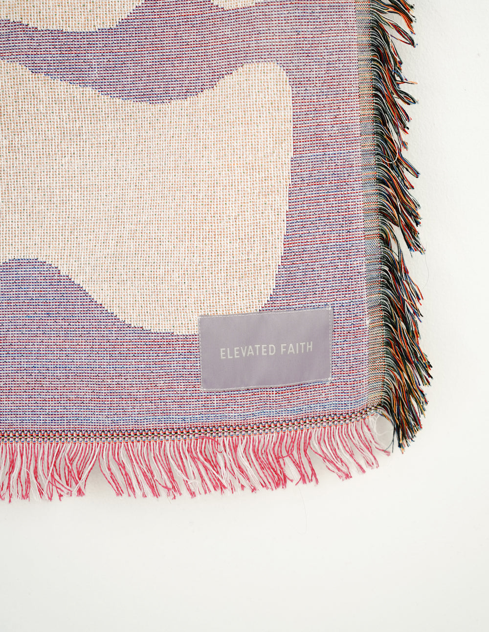 Throw blanket corner with woven fabric texture and 'Elevated Faith' patch, pink fringe