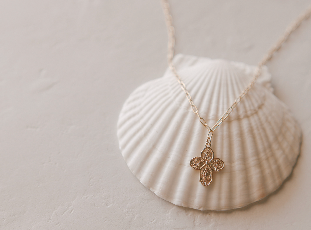 Gold cross pendant on paperclip chain necklace, laid over a shell