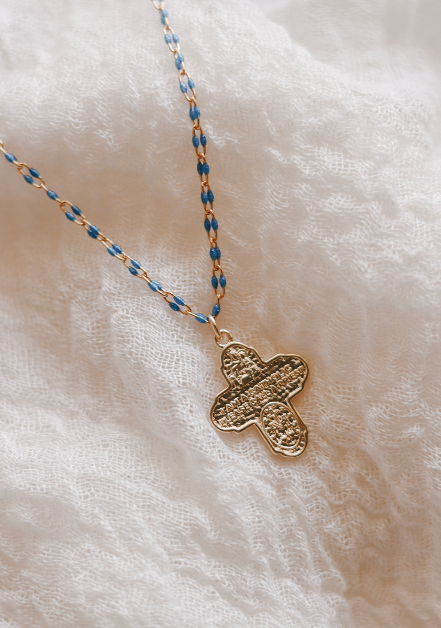Gold cross pendant necklace with blue beaded chain on light fabric.