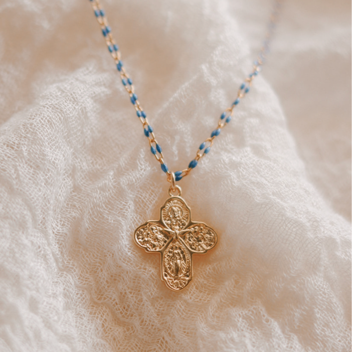 Gold four-way cross pendant necklace with blue beaded chain on textured fabric.