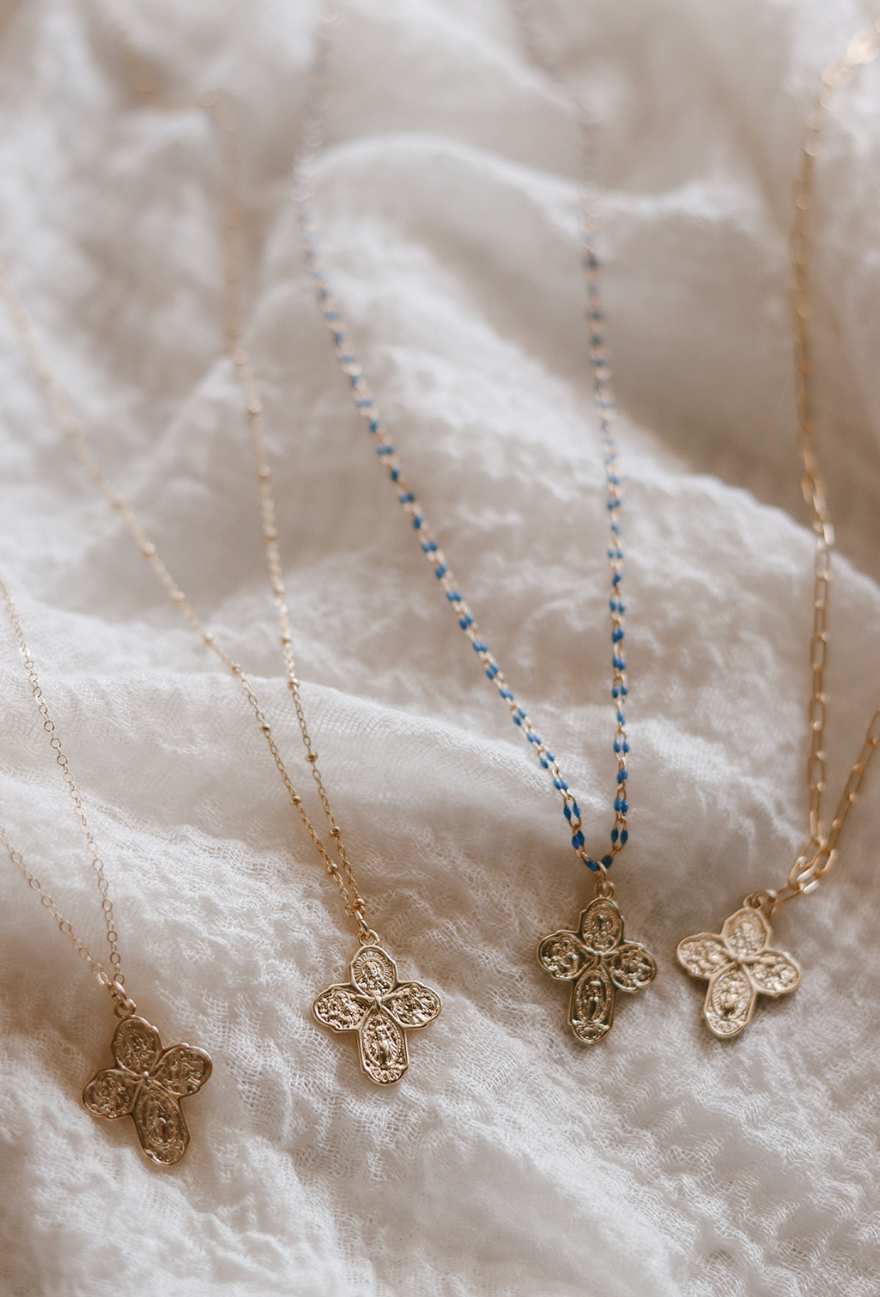 Four gold cross pendant necklaces with blue or gold chains laid on white fabric.