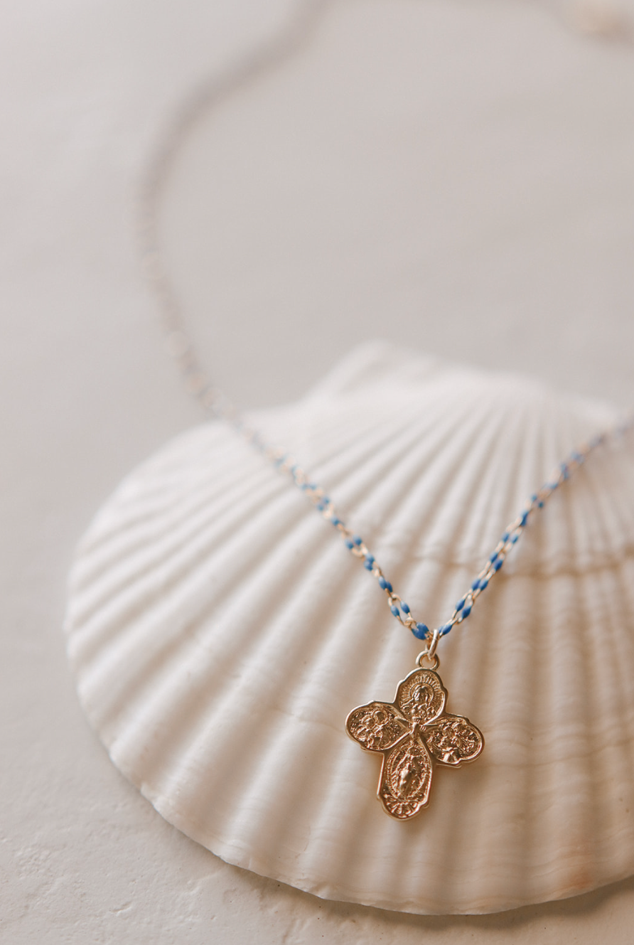 Gold four-way cross pendant necklace with blue beaded chain on white fabric.