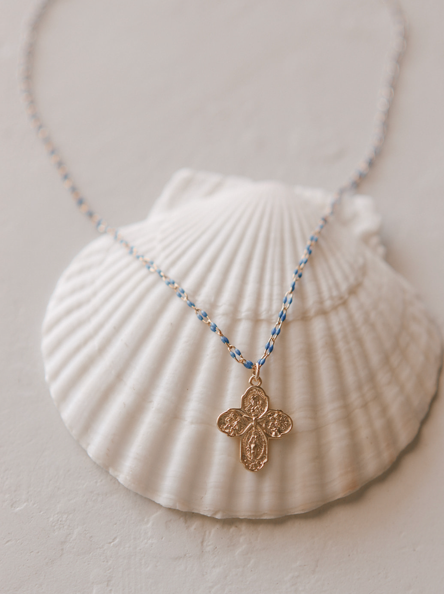 Gold cross pendant necklace with blue beaded chain on white shell pouch.