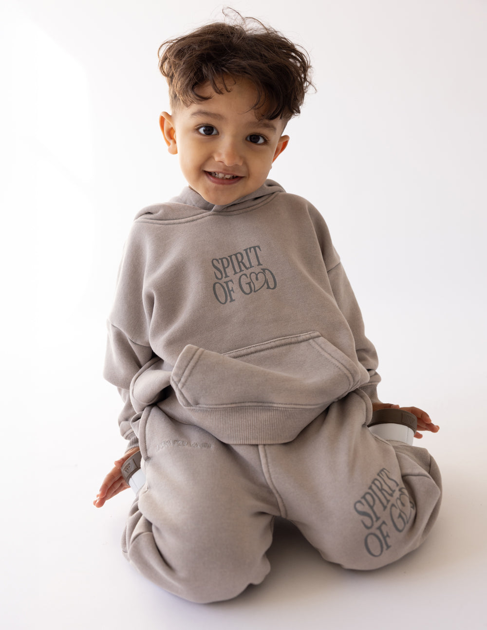 Kids taupe hoodie with hood, “SPIRIT OF GOD” chest print; child seated wearing it