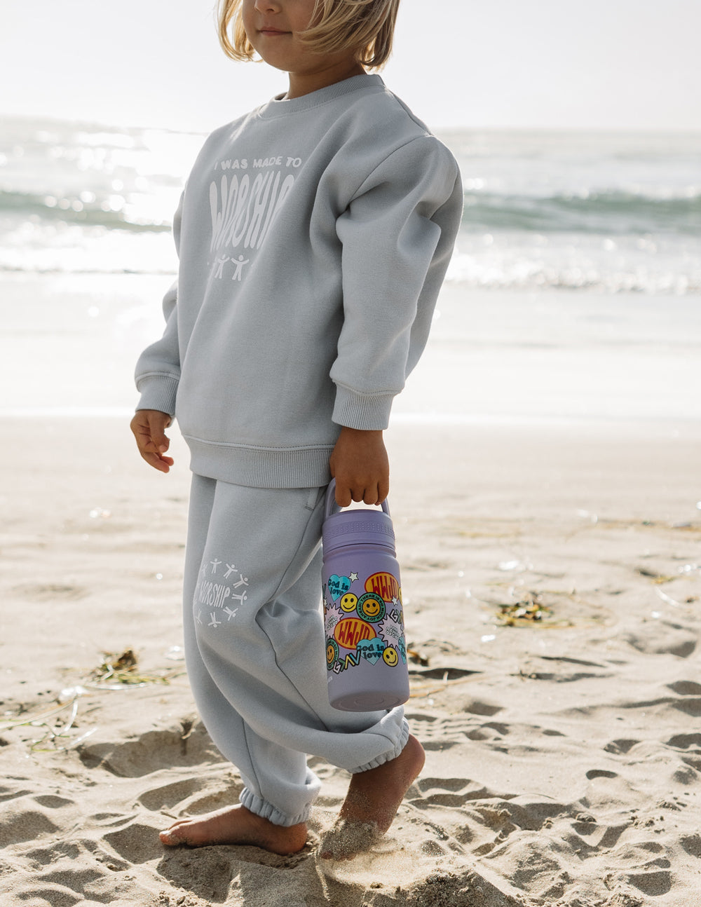 Lavender kids water bottle with colorful sticker collage, carried near beach sand