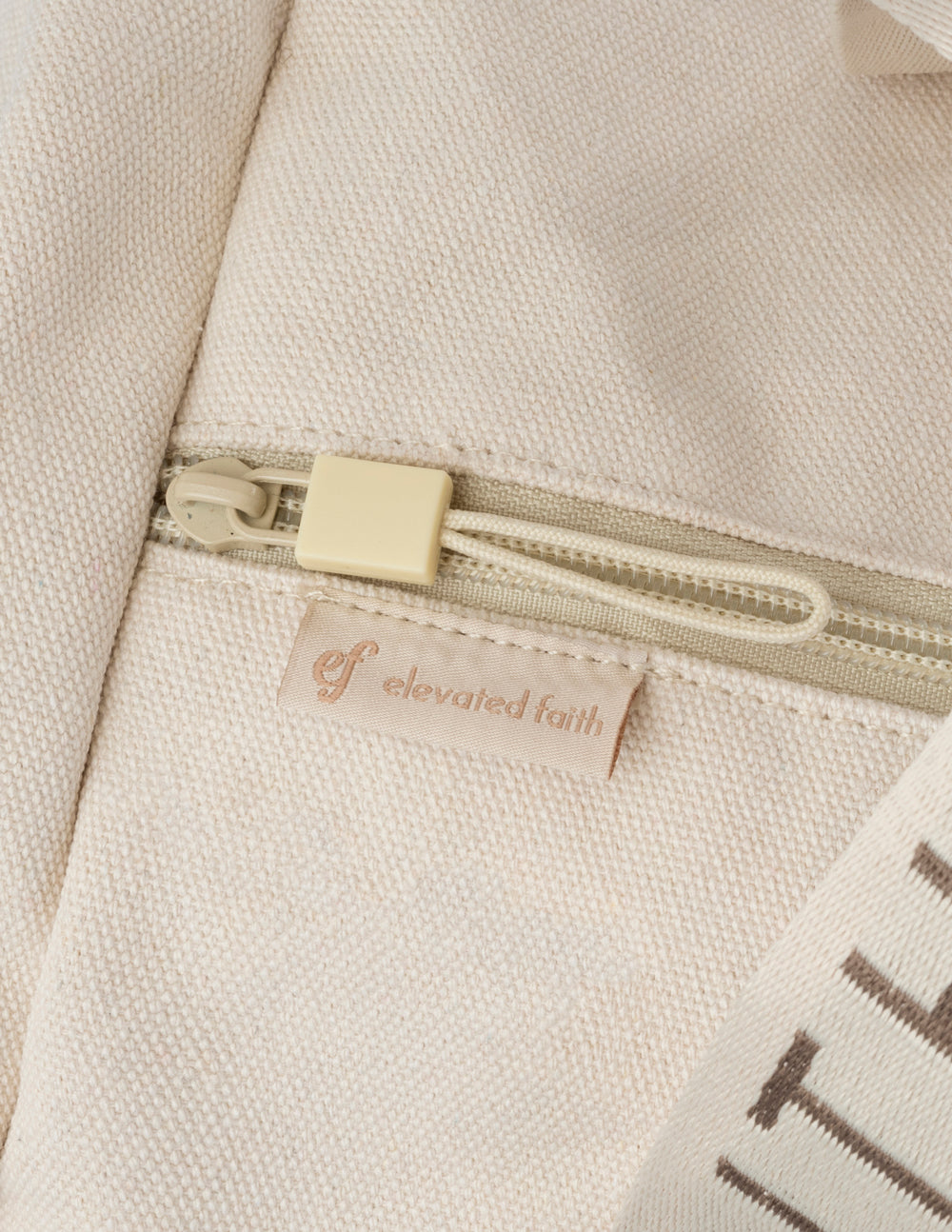 Cream canvas duffel bag zipper with Elevate Faith logo tab.