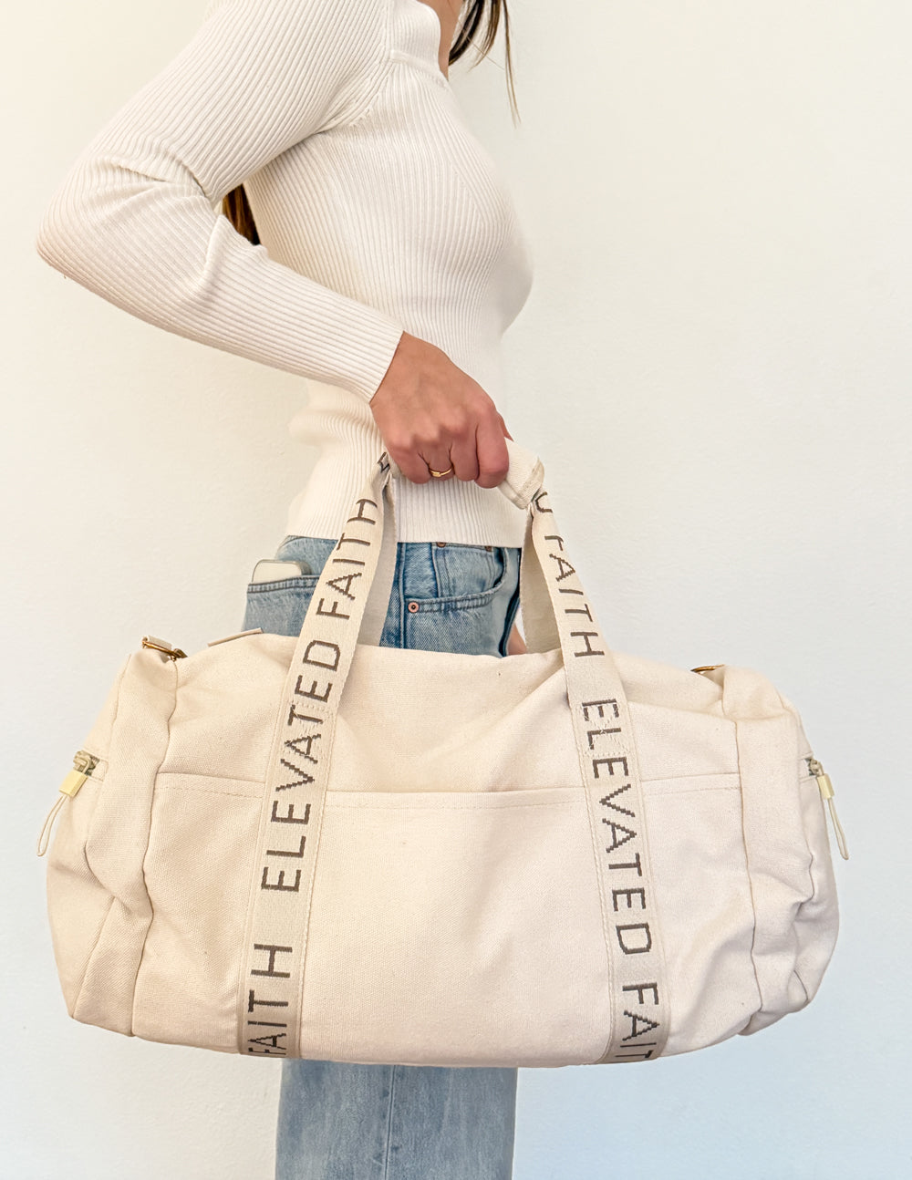 Cream duffel bag with Elevate Faith webbing straps and dual top handles.