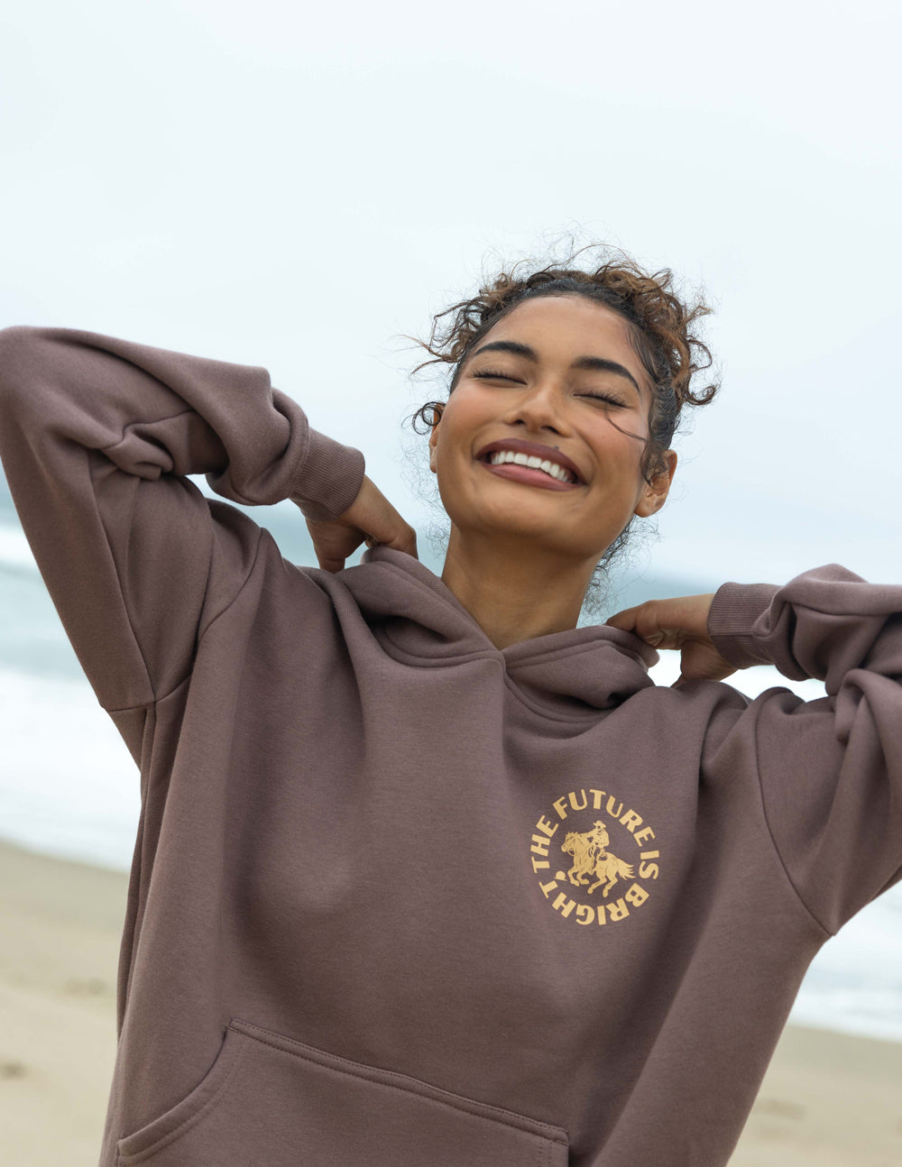 Mauve unisex hoodie with hood and gold chest graphic, worn with front pocket.