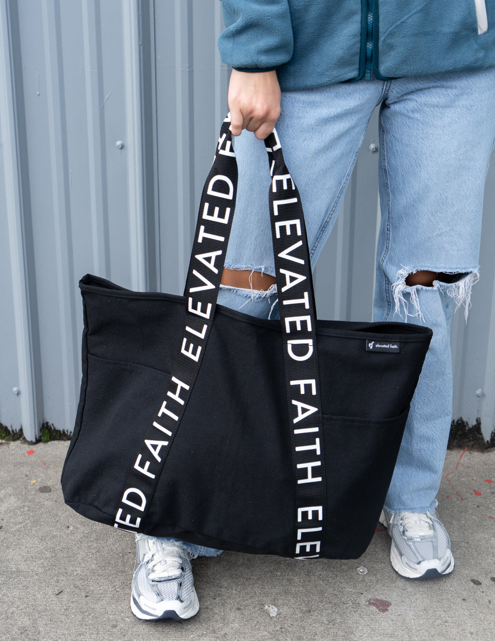 Black large tote bag standing with long handles and “ELEVATED FAITH” strap lettering