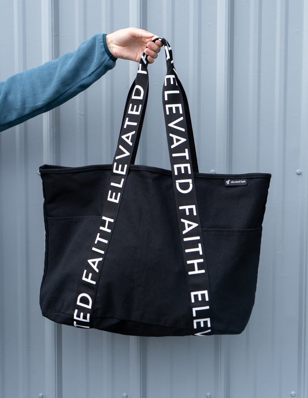 Black large tote bag held by one handle, showing “ELEVATED FAITH” vertical strap text