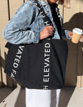 Black large tote bag with wide handles and vertical “ELEVATED FAITH” print on straps