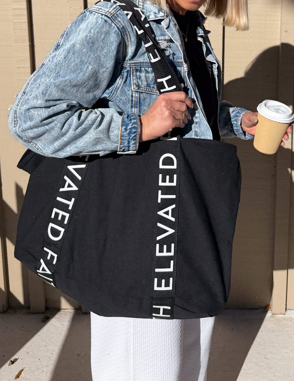 Black large tote bag with wide handles and vertical “ELEVATED FAITH” print on straps