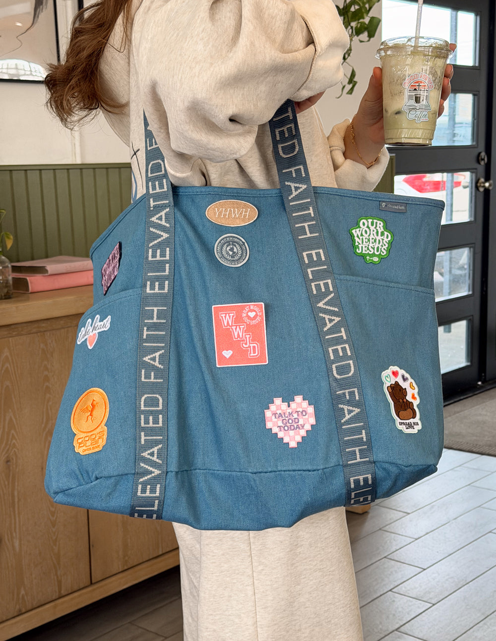 Blue denim tote bag with front patches and ELEVATED FAITH strap, held by person