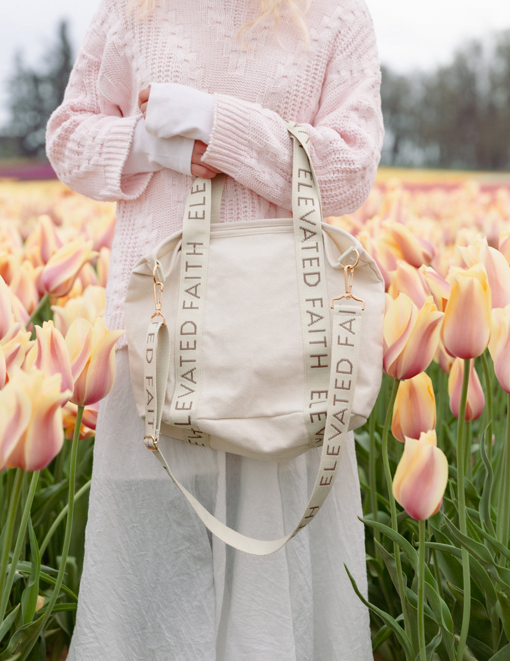 Cream mini tote held at the front with “ELEVATED FAITH” strap and gold-tone clasp