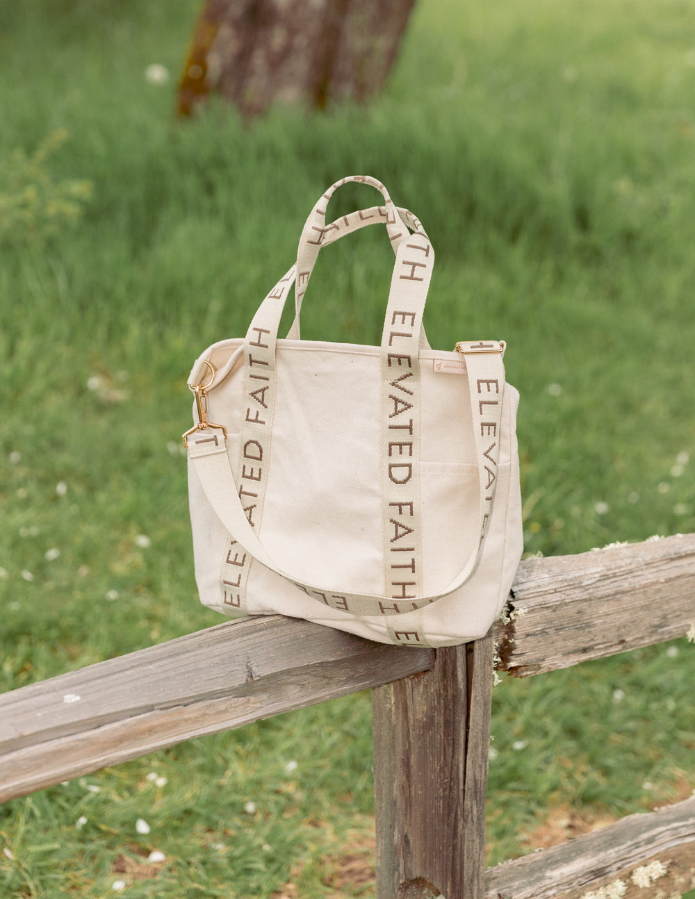 Cream mini tote on a wooden bench with “ELEVATED FAITH” strap and gold-tone handles