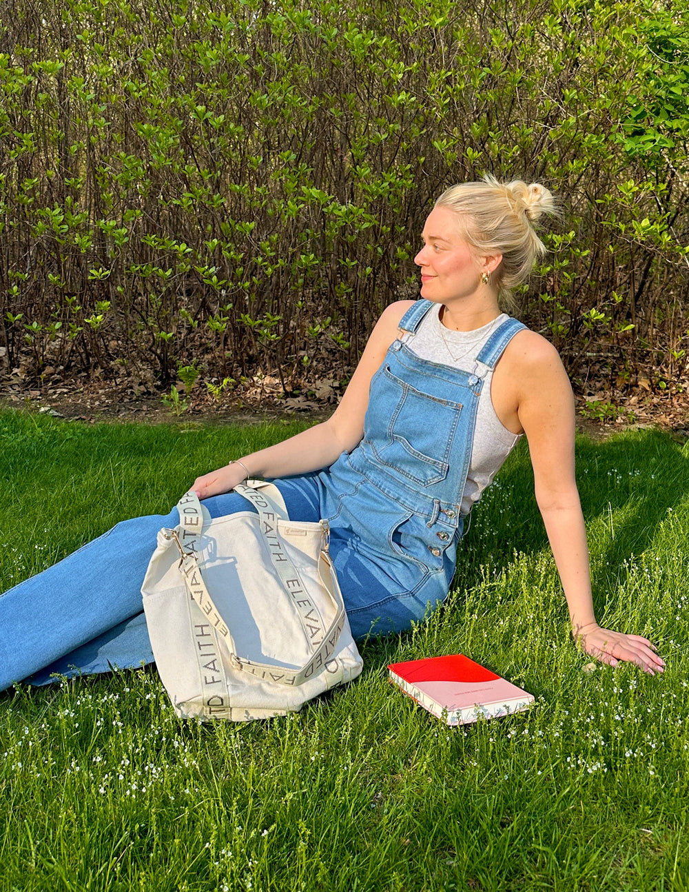 Cream mini tote bag with “ELEVATED FAITH” strap lettering beside the seated person