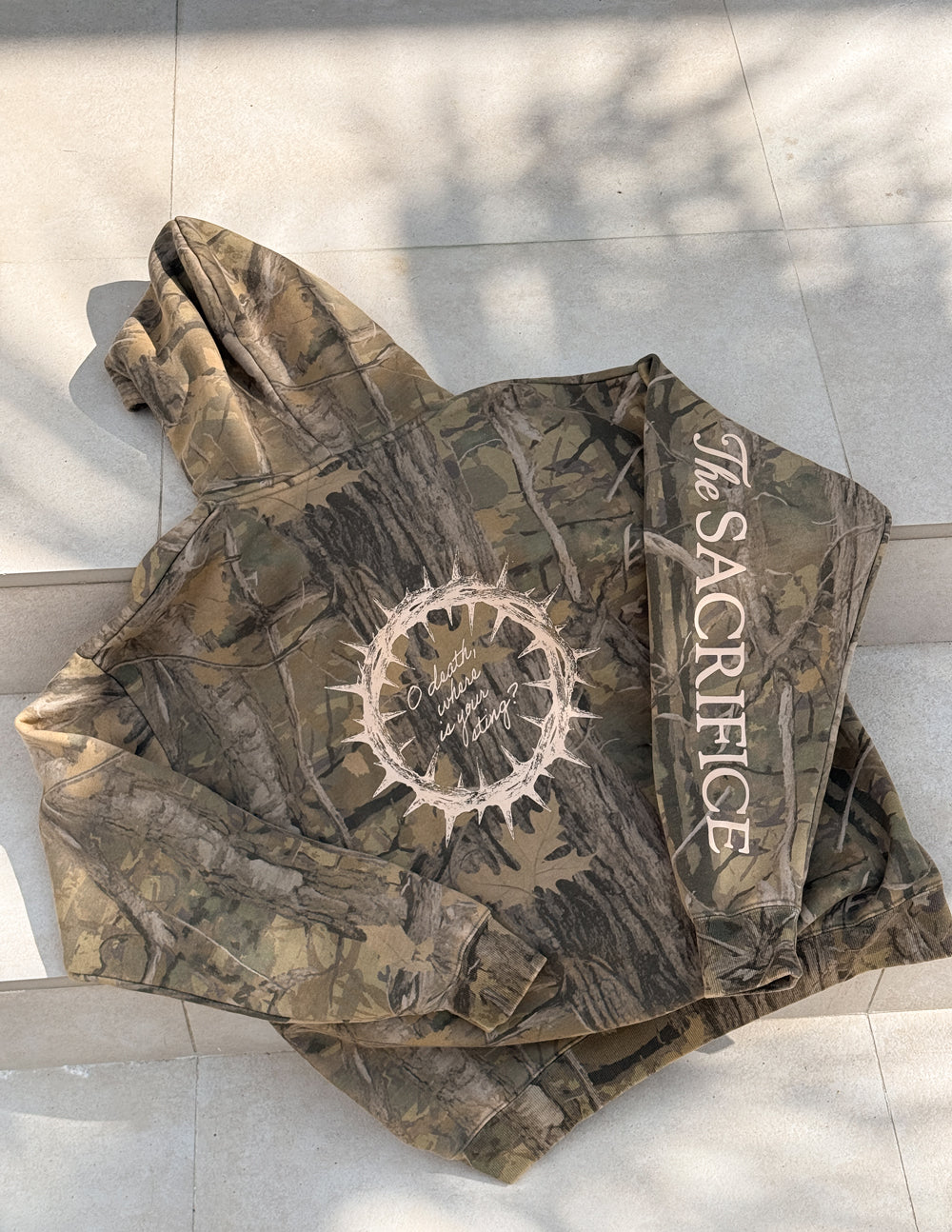 Camouflage unisex hoodie with bold circular center graphic and “Sacrifice” sleeve print