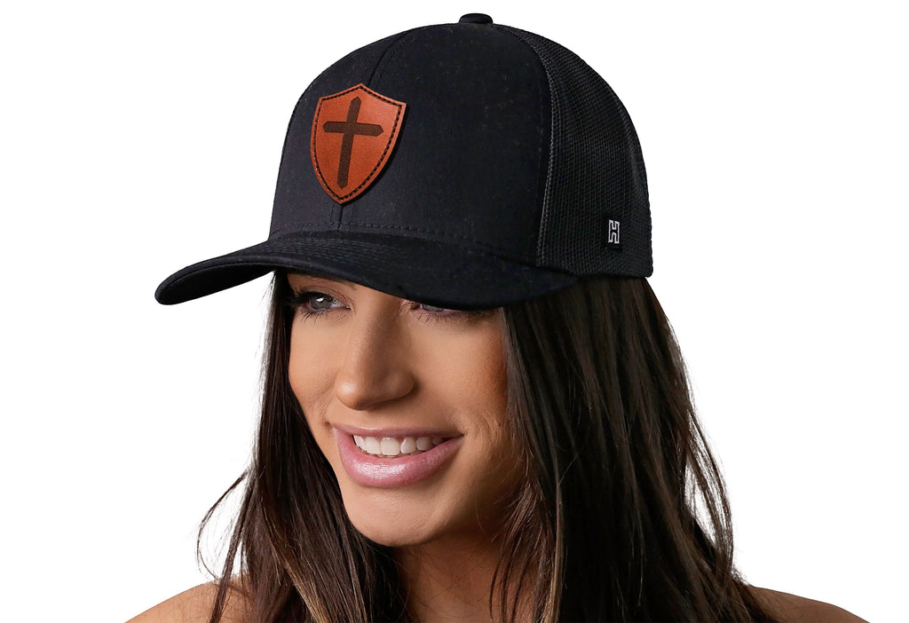 Black trucker snapback hat worn by woman, orange shield cross patch on front.
