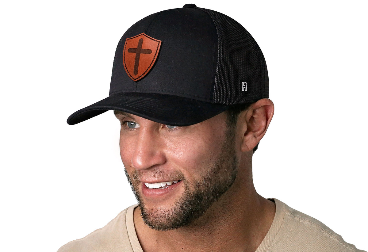 Black trucker snapback hat worn by man, orange shield cross patch on front.