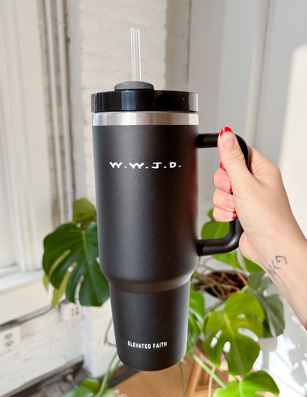 Black insulated tumbler with lid and straw, held by a hand with WWJD logo on front.