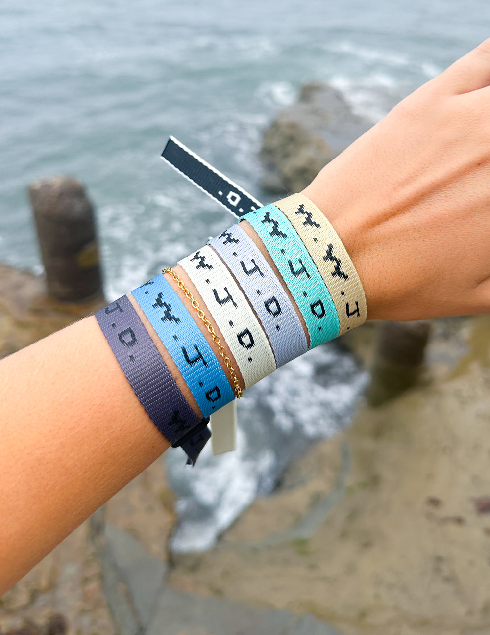 Stack of fabric surf bracelets with WWJD print on wrist, near ocean waves