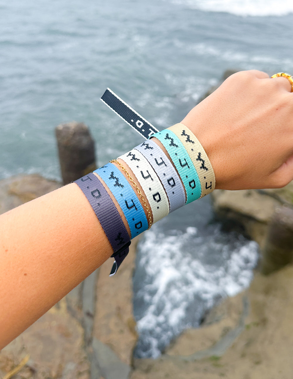 Stacked fabric surf bracelets with WWJD print on wrist, held at seaside rocks