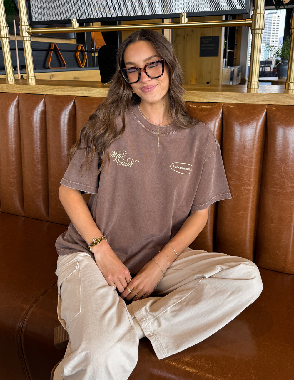 Brown crewneck T-shirt with 'Walk by Faith' logo and '2 Corinthians 5:7' oval front print.