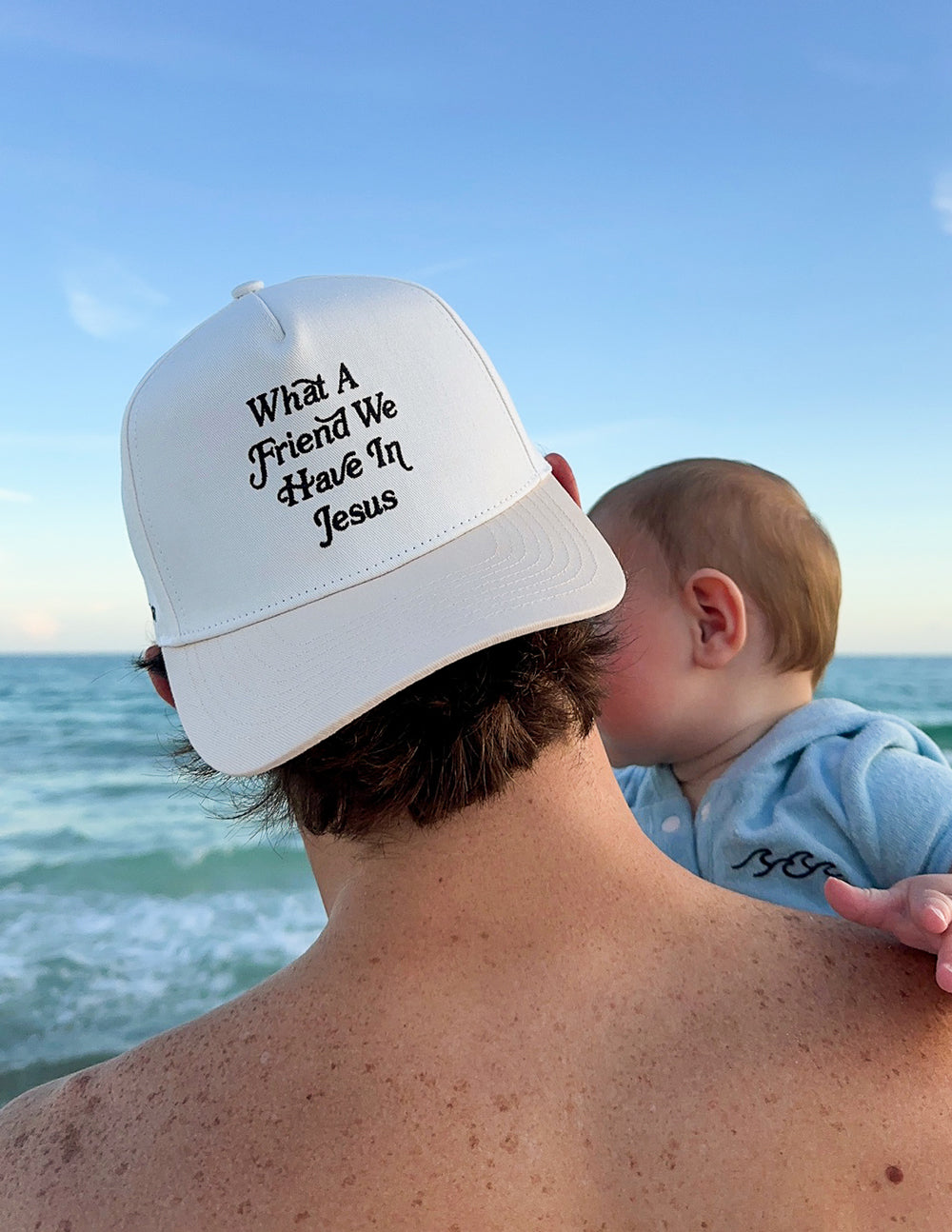 White cap with “What a Friend We Have In Jesus” embroidery, worn outdoors near sea