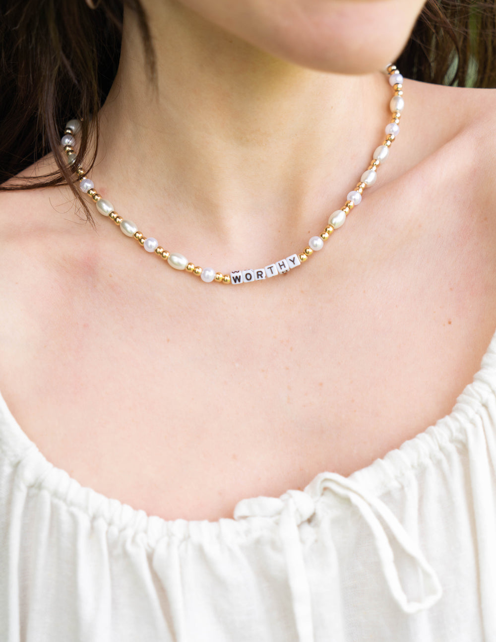 Letter necklace close-up with gold-tone beads, white pearls, and 'WORTHY' bar