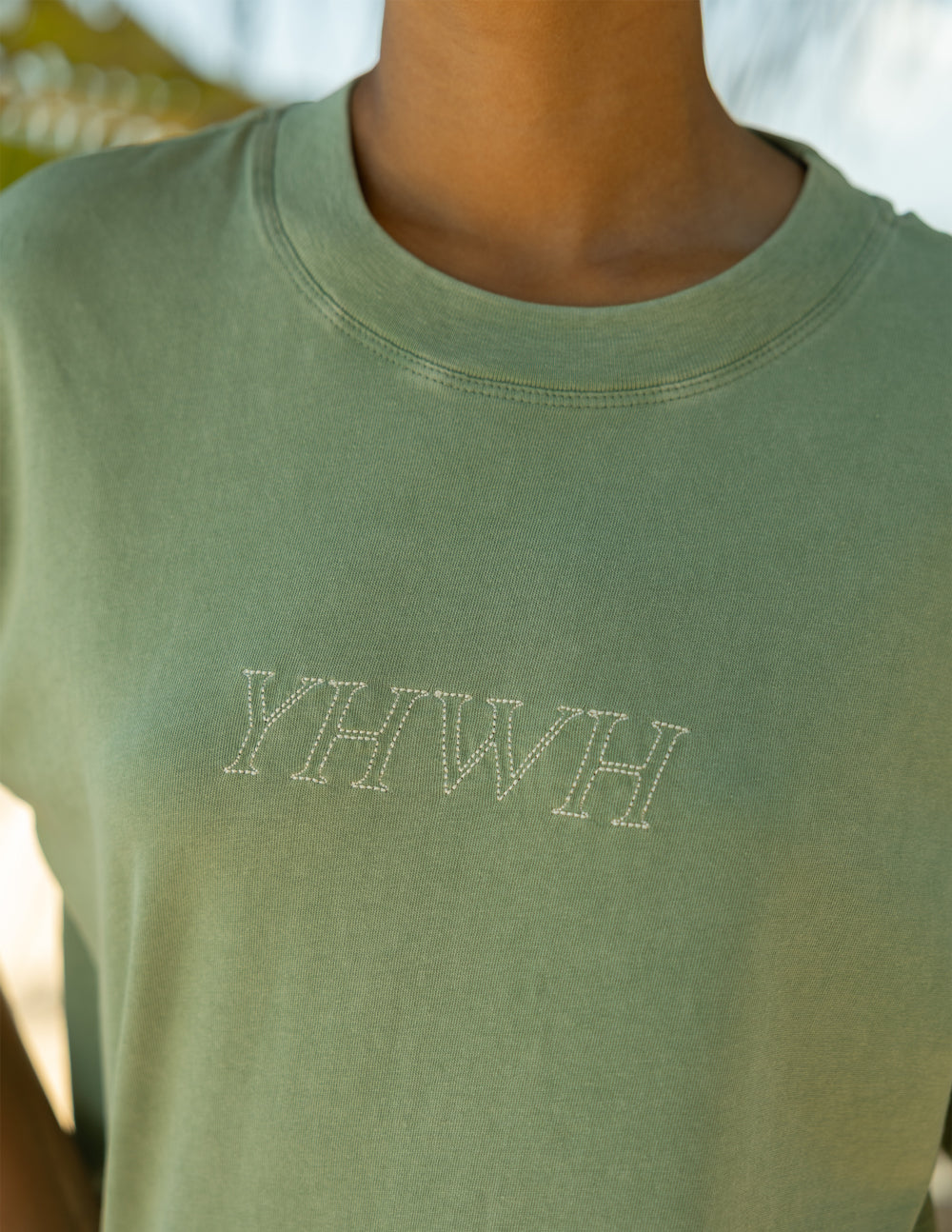 Sage green T-shirt close-up showing printed YHWH logo at center chest.