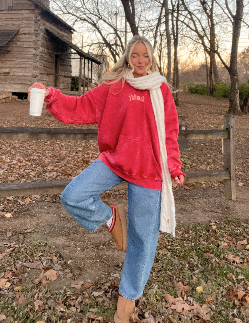 Red unisex hoodie with chest logo, worn with white scarf and jeans.