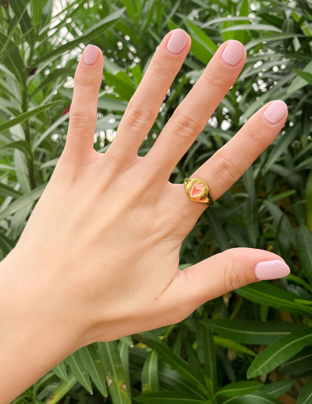 Gold-tone heart signet ring with green-pink heart emblem, worn on raised hand