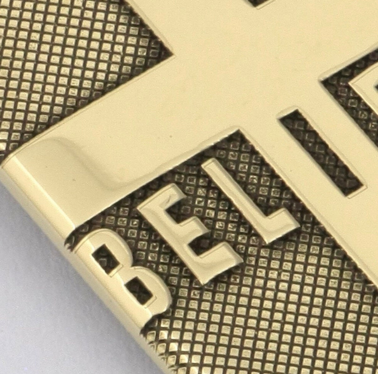 Gold-tone pendant close-up with 'BELIEVE' engraved letters over cross-hatch texture