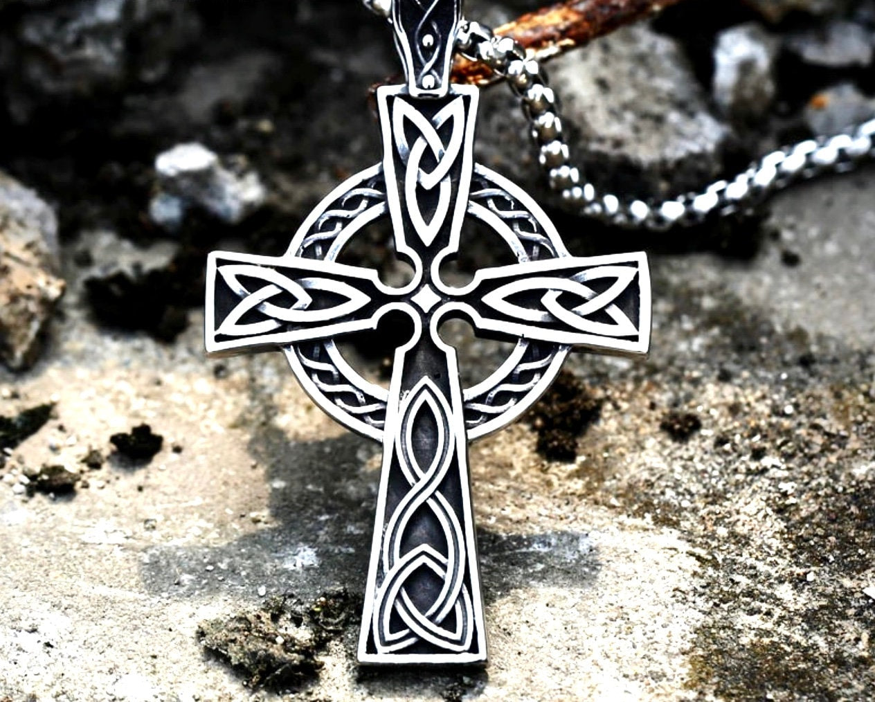 Silver Celtic knot cross pendant necklace on a chain, hanging over a rough stone surface