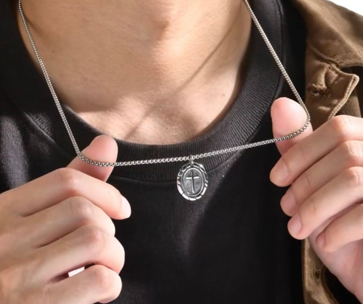 Silver-tone chain necklace with small oval cross pendant worn over dark shirt