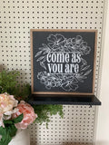 Framed floral wall sign with white script text reading “Come as you are”