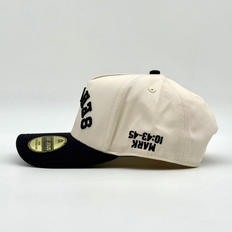 Cream snapback cap with black side lettering and logo patch, laid on side.