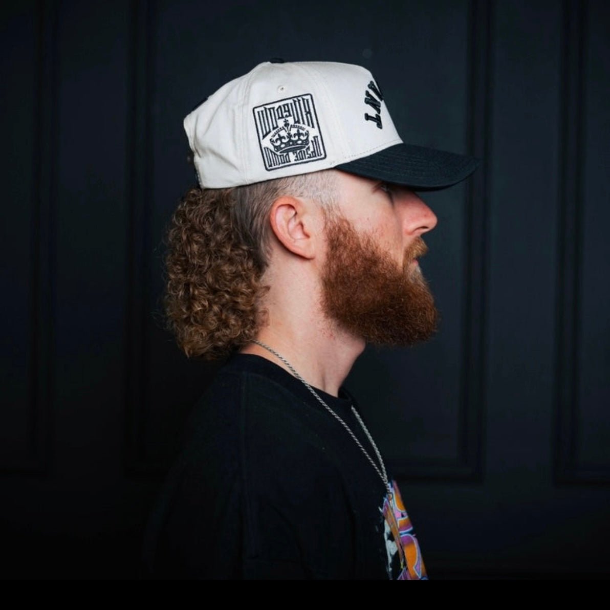 Cream snapback cap with black side and front prints, worn in profile.