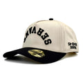 Cream snapback cap with black front lettering patch and dark brim.