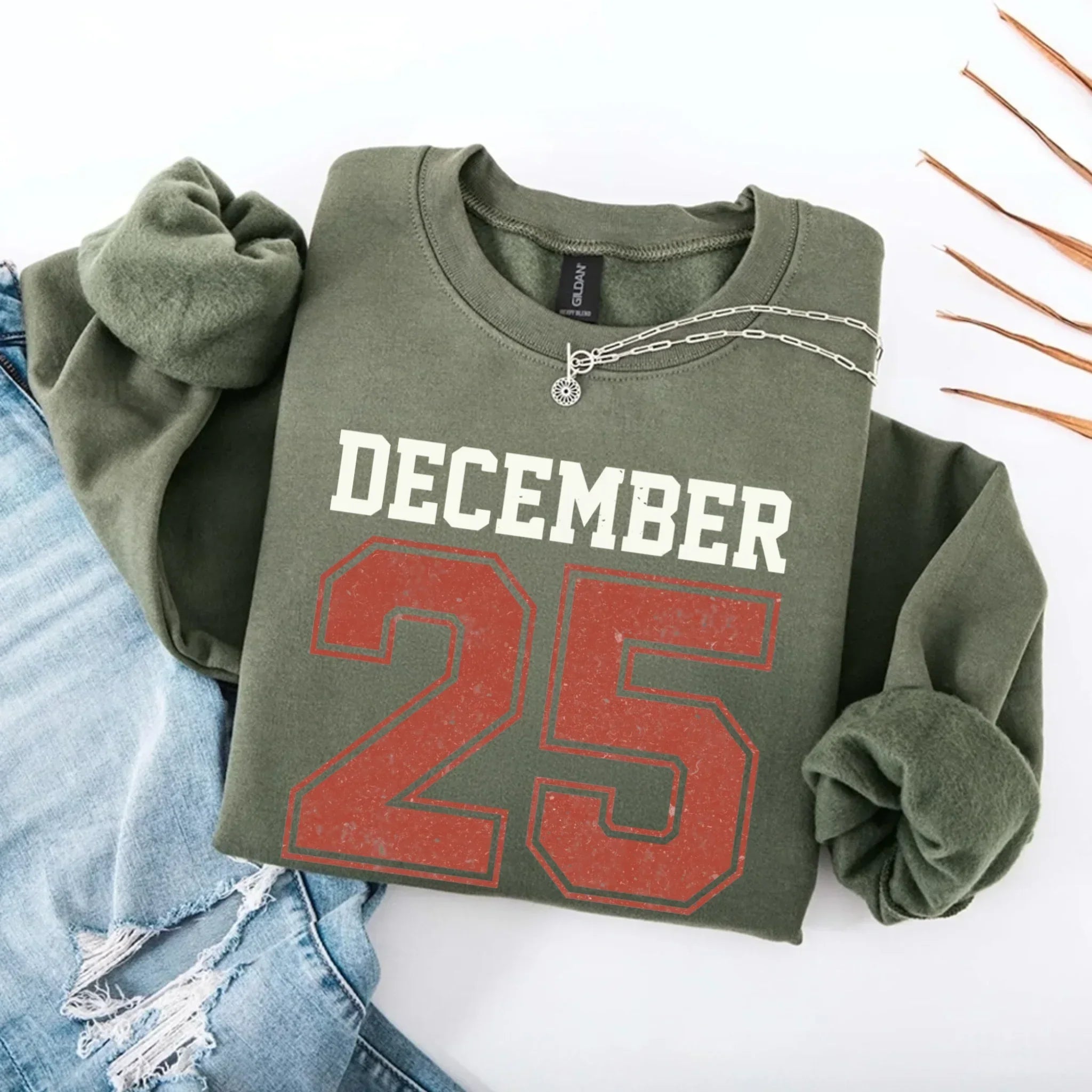 Olive green crewneck sweatshirt with “DECEMBER 25” graphic and silver chain detail.