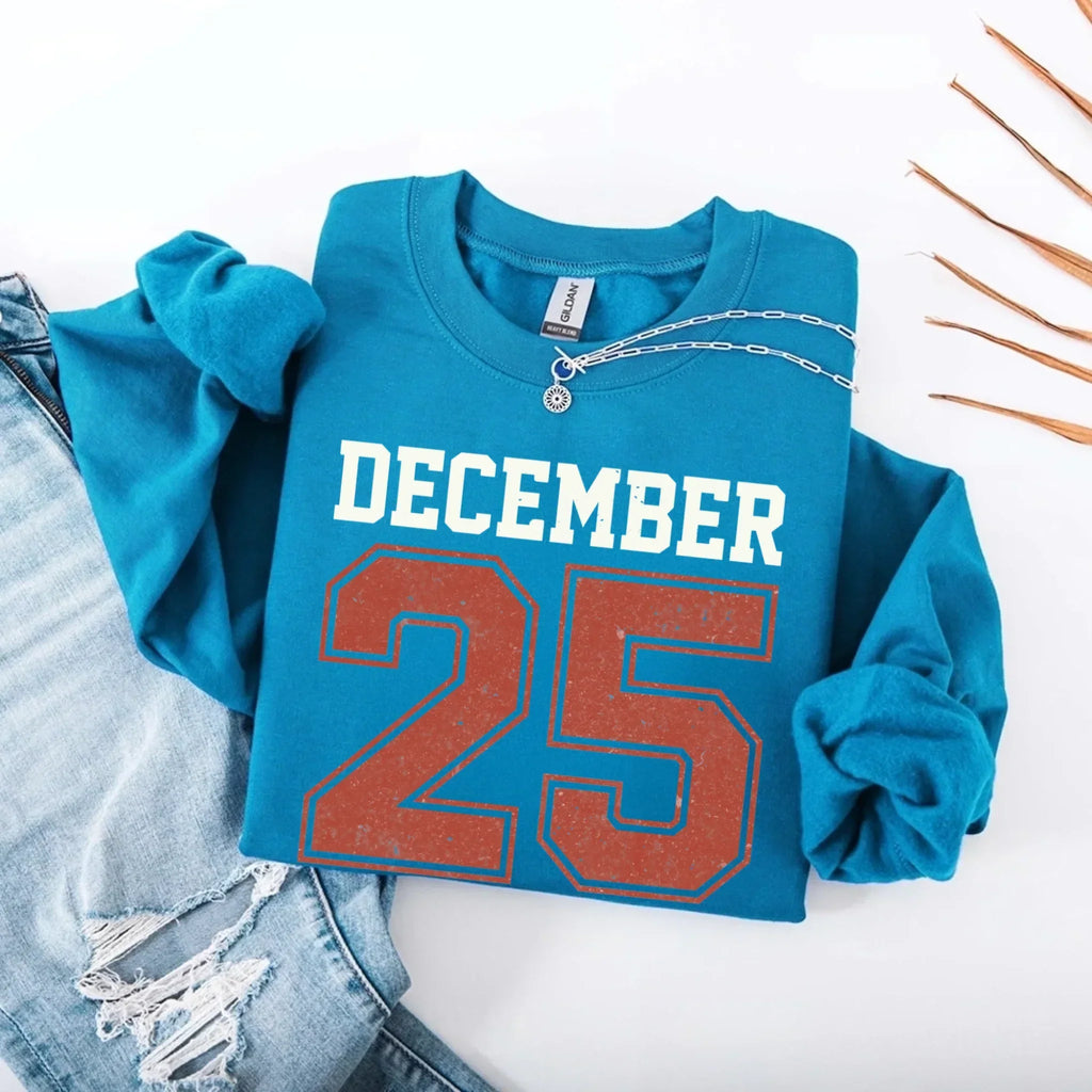 Blue crewneck sweatshirt with “DECEMBER 25” graphic and silver chain detail at collar.