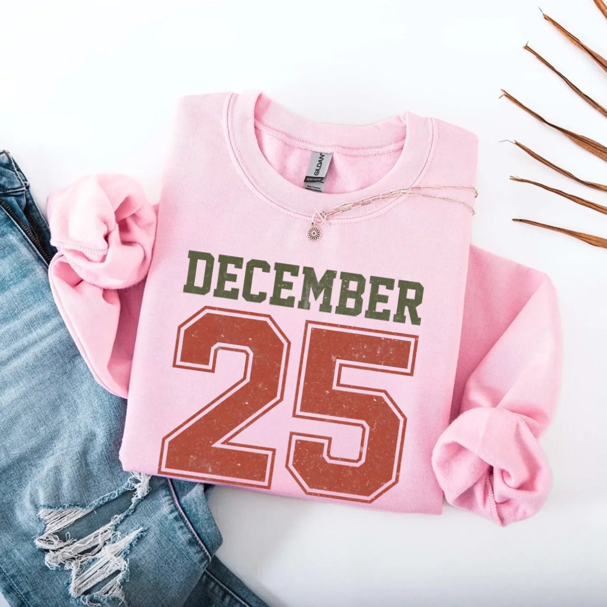 Pink crewneck sweatshirt with “DECEMBER 25” graphic and silver chain detail at neckline.