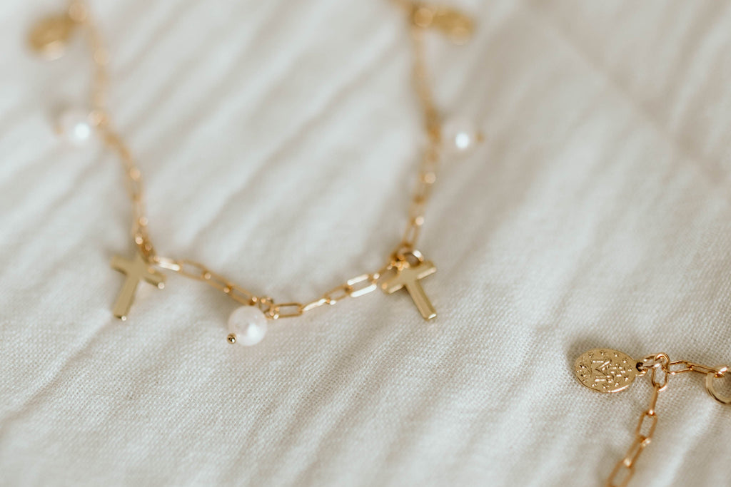 Gold-tone necklace on light fabric with cross charms and pearl center bead.