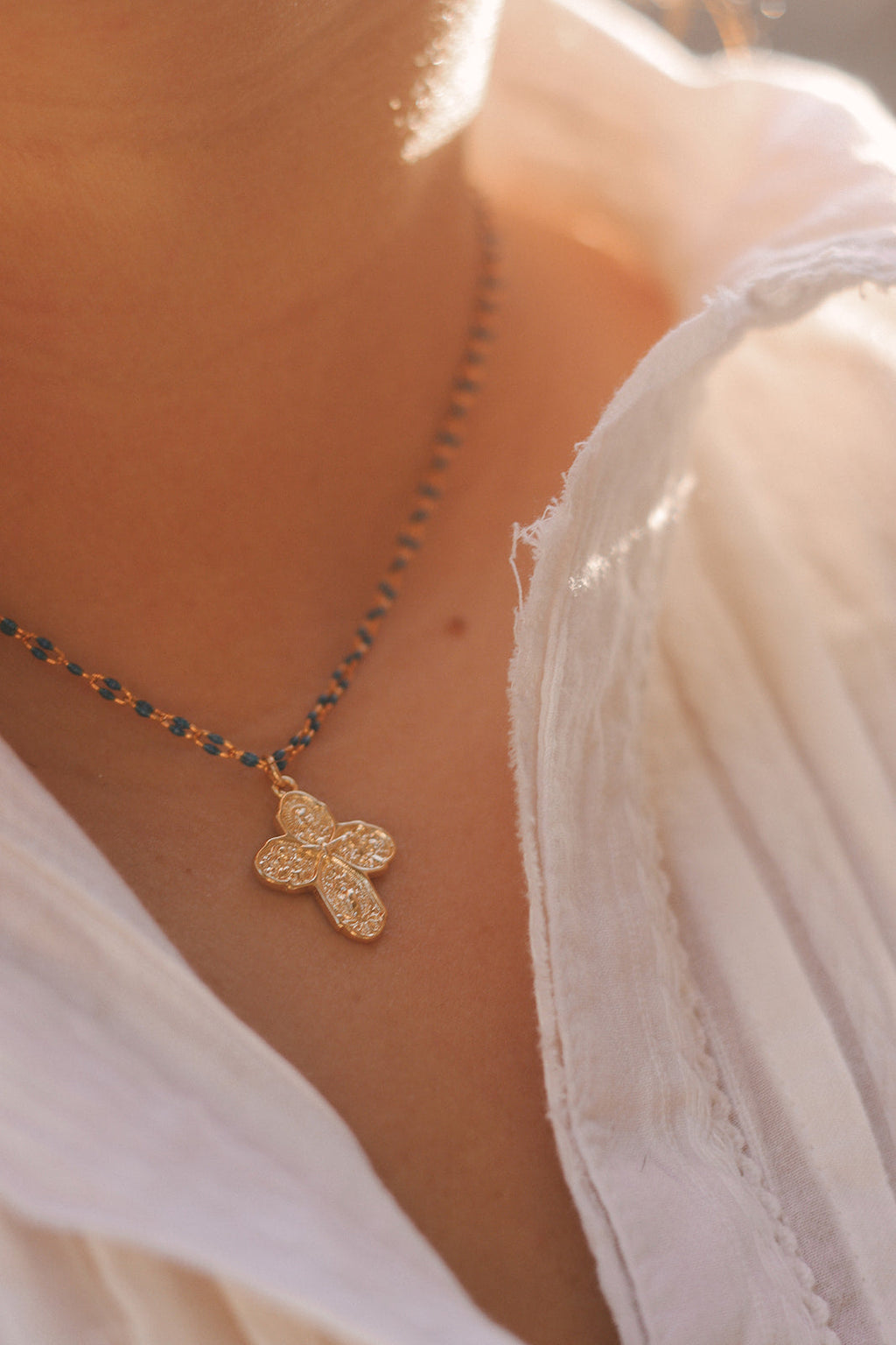 Gold cross pendant on blue beaded chain necklace against white dress fabric.