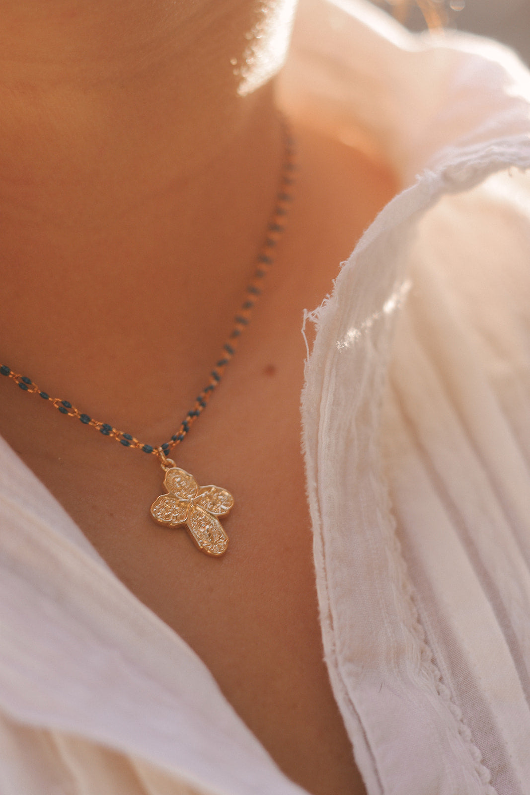 Gold cross pendant on blue beaded chain necklace against white dress fabric.